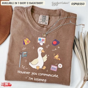 Puede incluir: Camiseta color espresso con un ganso de dibujos animados y el texto "However you communicate, I'm listening." La camiseta presenta varios iconos gráficos. Disponible en camiseta y sudadera.