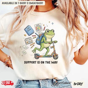 Può includere: T-shirt color avorio con un ranocchio dei cartoni animati su uno scooter, con uno zaino e materiale scolastico. Il testo "SUPPORT IS ON THE WAY" è stampato. Disponibile come t-shirt e felpa.