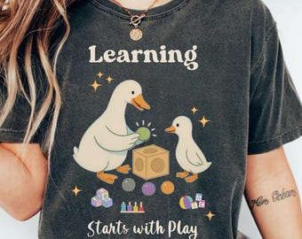 May include: Dark gray t-shirt with the words "Learning Starts with Play" and a graphic of two ducks with toys. The ducks are holding a green ball and standing near a wooden block, colorful balls, and toy blocks. The shirt is Comfort Colors brand.
