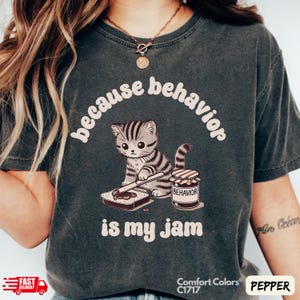 Puede incluir: Una camiseta gris oscura con el texto "because behavior is my jam" en blanco. Un gato de dibujos animados unta mermelada en una rebanada de pan. El tarro de mermelada dice "behavior".