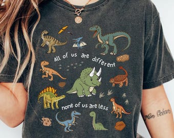 May include: Dark gray t-shirt featuring various cartoon dinosaurs in shades of green, brown, and blue. The text "All of us are different, none of us are less" is printed on the shirt. The shirt is labeled "Comfort Colors C1717 PEPPER".