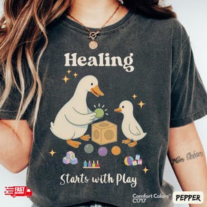 Op de afbeelding: Donkergrijs t-shirt met de tekst "Healing Starts with Play" en een afbeelding van twee eenden die met speelgoed spelen. Het shirt heeft ook het Comfort Colors-logo en de kleur "Pepper".