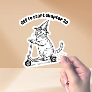May include: A black and white sticker featuring a cat wearing a witch hat and riding a scooter. The text "Off to start chapter 30" is above the cat. The sticker is held by a hand.
