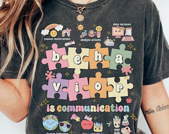 Op de afbeelding: Een donkergrijs T-shirt met een kleurrijk grafisch ontwerp van puzzelstukjes die "gedrag is communicatie" spellen. De grafiek bevat illustraties van een regenboog, een bloem, een potlood, een penseel en een wereldbol. De tekst "Comfort Colors C1717 PEPPER" staat gedrukt op de onderkant van het shirt.
