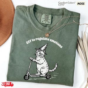 May include: Moss green Comfort Colors C1717 t-shirt with a graphic of a cat wearing a witch hat riding a scooter. The text "Off to regulate emotions" is above the cat. A gold necklace is on the shirt. The shirt has a Comfort Colors tag.