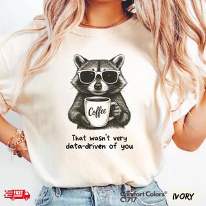 May include: Ivory-colored t-shirt featuring a black and white illustration of a raccoon wearing sunglasses and holding a coffee mug. The mug has the word "Coffee" printed on it. Below the raccoon is the text "That wasn't very data-driven of you."