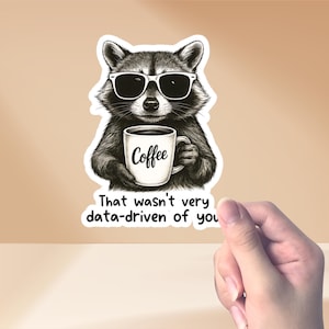 Op de afbeelding: Een sticker met een wasbeer met een zonnebril die een koffiemok vasthoudt. Op de mok staat "Coffee". Onder de wasbeer staat de tekst "That wasn't very data-driven of you."
