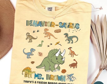 May include: A butter yellow t-shirt featuring the text "BEHAVIOR-SAURUS" and "MS. BROWN There's a reason BeHind every roar" with various colorful dinosaur illustrations including a large green triceratops, footprints, and small fossils.