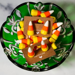 May include: A pair of handmade earrings featuring a candy corn design. The earrings are displayed on a green and white patterned surface with the words "Handmade" and "and made" on the cards.