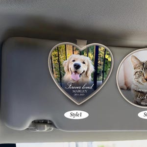 May include: Two car visor memorial ornaments. The first is a heart-shaped ornament with a photo of a golden retriever and the text "Forever loved Marley 2011-2023". The second is a round ornament with a photo of a tabby cat and the text "Forever loved Lucky 2024".