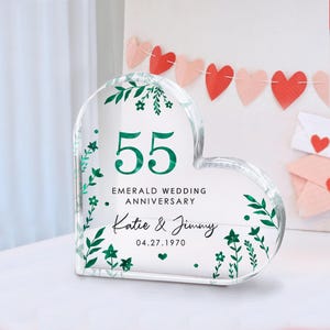 Peut inclure: Un souvenir en acrylique transparent en forme de cœur célébrant le 55e anniversaire de mariage émeraude. Le design présente le chiffre "55" en vert, les noms "Katie & Jimmy" et la date "04.27.1970". Des motifs floraux verts et du texte sont inclus.
