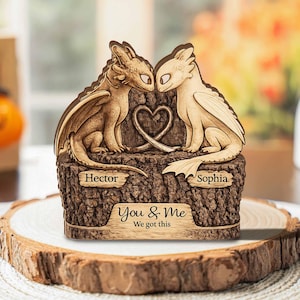 May include: A wooden sculpture featuring two dragon figures facing each other, with a heart-shaped design between them. The base is engraved with the names "Hector" and "Sophia", and the text "You & Me We got this". The sculpture is made of light brown wood.