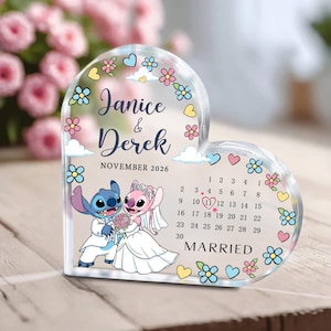 Peut inclure: Un souvenir de mariage en acrylique en forme de cœur avec un couple de dessins animés, Stitch et Angel, en tenue de mariage. Les noms "Janice & Derek" et la date "Novembre 2026" sont affichés, avec un calendrier marquant la date du mariage. Des motifs floraux et de nuages décorent les bords.