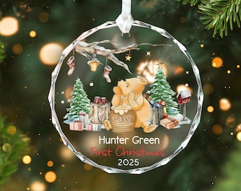 Op de afbeelding: Een heldere, facetgeslepen kerstbal met een Winnie de Poeh-ontwerp. De ornamenten hebben een beer, een honingpot, kerstbomen en de tekst "Hunter Green First Christmas 2025". Het hangt aan een wit lint.