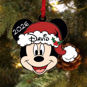 Könnte beinhalten: Ein weihnachtlicher Baumschmuck in Form von Mickey Mouse mit einer roten Weihnachtsmütze mit weißem Rand, einem Stechpalmenzweig, dem in Schwarz gedruckten Namen David und der Jahreszahl 2026. Der Schmuck hängt an einem dünnen roten Band.