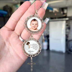 Op de afbeelding: Een zilverkleurige hangerketting met twee sierlijke, cirkelvormige bedels. De bovenste bedel bevat een babyfoto, terwijl de onderste bedel een afbeelding van Jezus en een citaat heeft. Een klein kruisje hangt eronder. De tekst luidt "Baby Brown 05.10.28".
