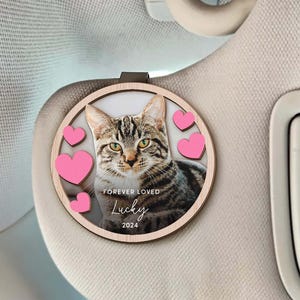 May include: A wooden car charm with a photo of a tabby cat and pink hearts. The charm has the text "Forever Loved Lucky 2024" on it.