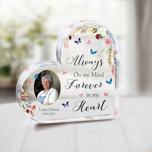 Pode incluir: Uma lembrança memorial em forma de coração com uma foto de uma mulher, Lucy Green, e os anos 1950-2023. A peça de acrílico transparente tem detalhes florais e borboletas e as palavras "Always On my Mind Forever in my Heart."