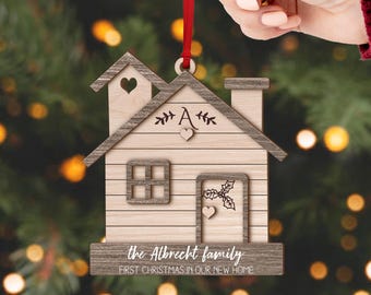 Puede incluir: Adorno navideño de madera con forma de casa y cinta roja. El adorno presenta un recorte en forma de corazón, una chimenea y una puerta con un diseño de acebo. El texto "the Albrecht family FIRST CHRISTMAS IN OUR NEW HOME" está grabado en la parte inferior.