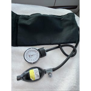 Puede incluir: Un manguito de presión arterial negro con un medidor de esfera blanca y una pera negra. El medidor tiene una esfera circular con números y una aguja. La pera tiene una etiqueta amarilla con texto. El manguito está sobre una superficie blanca.