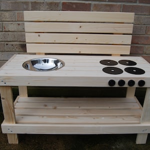 May include: A wooden play kitchen in a natural finish. It includes a stainless steel bowl, four black burner rings, and four black knobs. The kitchen has a backboard and a lower shelf. The play kitchen is designed for children to play with.