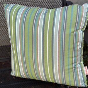 May include: Square outdoor pillow with a green and white striped pattern. The pillow has vertical stripes in shades of green, white, gray, and blue. The pillow is resting on a black metal surface.