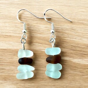 May include: Dangle earrings with silver hooks, each with three light blue and one brown sea glass pieces. The sea glass is linked with silver-coloured metal. The earrings are shown on a wooden surface.
