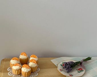Peut inclure: Une table en bois miniature avec une dentelle blanche et une assiette de cupcakes surmontés d'oranges. Une petite assiette avec un bouquet de fleurs de lavande se trouve sur une nappe à carreaux bleus et blancs.