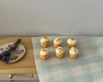Peut inclure: Six mini cupcakes au gâteau de carotte avec glaçage blanc et décorations de carottes orange sur une nappe à carreaux bleu clair et blanc.