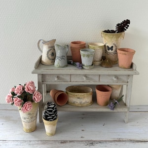 May include: A miniature potting bench displays a collection of small terracotta pots, decorative pitchers, and a vase with pink roses. The bench also holds a pinecone and lavender sprigs, creating a charming, rustic scene.
