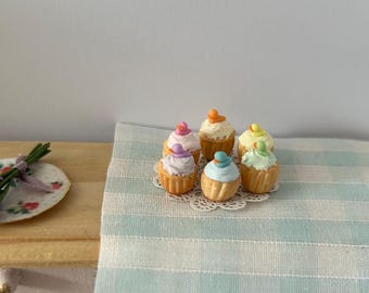 Peut inclure: Une collection de six mini cupcakes avec un glaçage de couleur pastel et des décorations colorées sur le dessus. Les cupcakes sont disposés sur un napperon blanc sur une nappe à carreaux bleu clair et blanc.