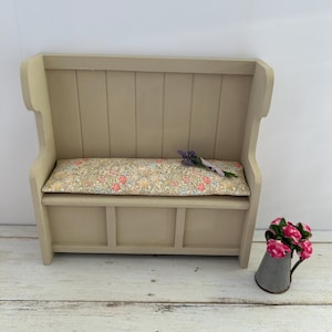 May include: A light beige wooden bench with a floral patterned cushion and a small metal watering can with pink flowers. The bench has a high back and a storage compartment underneath.