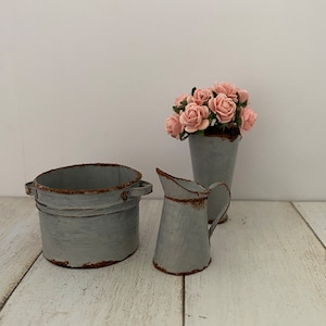 May include: Three gray metal containers with a rustic finish. The largest container is a round pot, the middle container is a vase with a bouquet of pink paper flowers, and the smallest container is a pitcher.