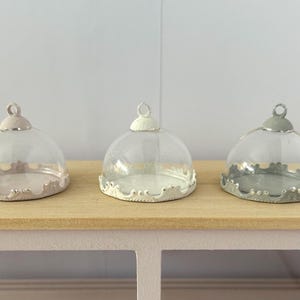 May include: Three glass cloches with decorative, scalloped edges and crown-shaped bases. Each cloche has a different pastel colour on the top and base: pink, white, and grey. The cloches are displayed on a light wood surface.