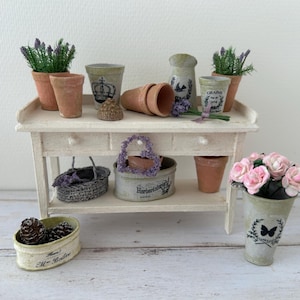 Puede incluir: Una mesa de jardinería blanca en miniatura exhibe macetas de terracota, plantas de lavanda y una cesta. Un pequeño recipiente contiene piñas, y un jarrón con flores rosas está a la derecha. La mesa tiene dos cajones y un estante inferior.