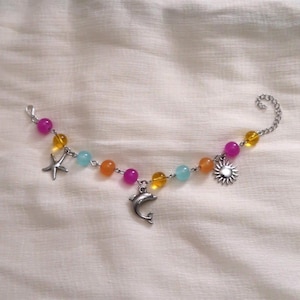 May include: A colorful beaded bracelet with a silver chain and charms. The bracelet features a starfish, dolphin, and sun charm, along with beads in shades of pink, yellow, orange, and blue. The bracelet is designed for a beach or summer theme.