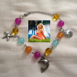 May include: A beaded bracelet with a silver chain and charms. The bracelet features colorful beads in shades of purple, yellow, orange, and blue. Charms include a starfish, a sun, and a dolphin. An image of a woman in an orange dress is in the center.