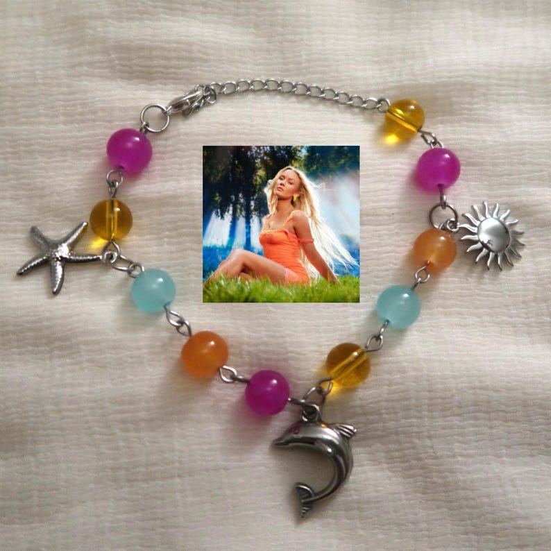 May include: A beaded bracelet with a silver chain and charms. The bracelet features colorful beads in shades of purple, yellow, orange, and blue. Charms include a starfish, a sun, and a dolphin. An image of a woman in an orange dress is in the center.