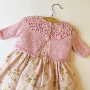 Puede incluir: Un cárdigan de punto rosa con un delicado patrón de encaje y cierre de botón, combinado con un vestido de estampado floral.