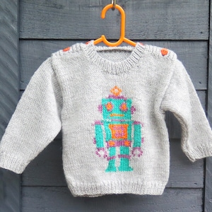 May include: A gray knitted sweater with a robot design in teal, orange, pink, and purple. The robot has a small antenna on its head and is standing with its arms at its sides.