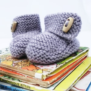 May include: A pair of lavender knitted baby booties with wooden button closures. The booties are sitting on a stack of children's books.