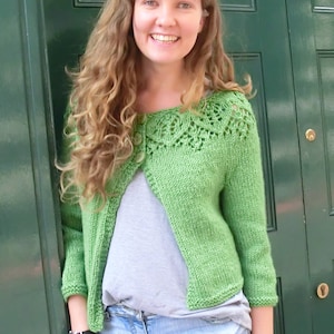 May include: A light green knitted cardigan with a decorative openwork neckline and three buttons. The cardigan is paired with a gray top and denim shorts. The person is wearing a watch and bracelets.