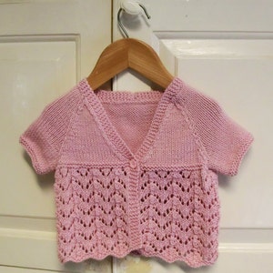 May include: A pink knitted cardigan with short sleeves and a button closure. The cardigan has a delicate lace pattern and a scalloped hem.