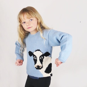 May include: A young girl wearing a light blue knitted sweater with a black and white cow design. The sweater has long sleeves and a crew neck.