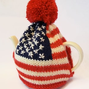 Puede incluir: Un calienta-tetera de punto rojo, blanco y azul con un pompón rojo en la parte superior. El calienta-tetera está diseñado para parecerse a la bandera estadounidense.