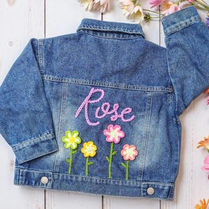 May include: A blue denim jacket with the name "Rose" embroidered on the back in pink thread. The jacket has four embroidered flowers with yellow, pink, and white petals.