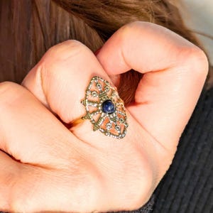 Könnte beinhalten: Ein goldener Ring mit einem filigranen, komplizierten Design, das einen blauen Edelstein in der Mitte trägt.