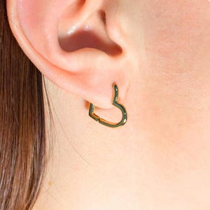 Pode incluir: Brincos de argola de coração de ouro. Os brincos são usados na orelha de uma pessoa.
