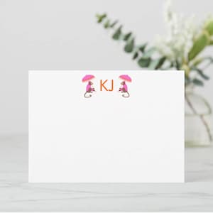 May include: White stationery card with two monkeys wearing pink umbrellas and holding a stick. The monkeys are facing each other and the initials "KJ" are printed in orange in the center.