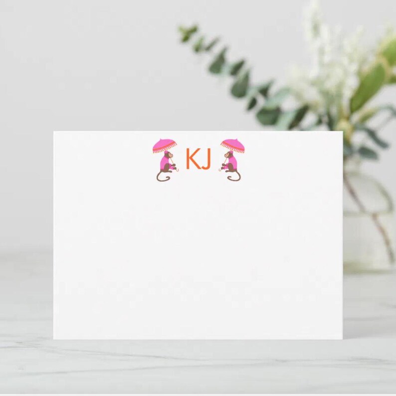 May include: White stationery card with two monkeys wearing pink umbrellas and holding a stick. The monkeys are facing each other and the initials "KJ" are printed in orange in the center.